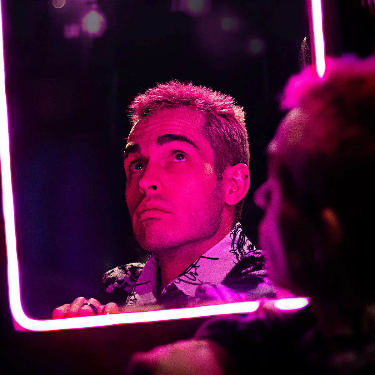 Corné Prinsloo looking up into a mirror framed by pink neon light, his reflection lit magenta, with the back of his head blurred in the foreground.